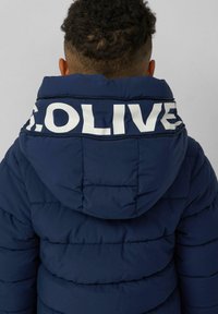 Kind in een marineblauwe gewatteerde jas met capuchon, met de rug naar de camera, gedeeltelijke witte tekst "OLIVE" zichtbaar op de capuchon.