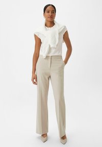 Un pantalon beige ajusté associé à un haut blanc et un pull en tricot drapé sur les épaules. Un tissu texturé et des escarpins à bout pointu complètent le look.