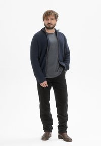 Marineblauwe cardigan met ribbeltextuur, ritssluiting aan de voorkant, gedragen over een grijs t-shirt, gecombineerd met zwarte broek en bruine laarzen. Simpel, casual ontwerp.