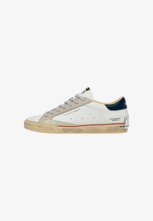 Sneaker bianca low-top con area delle stringhe in suede beige, linguetta del tallone blu navy, striscia rossa sulla suola in gomma sporca e testo laterale dorato "Céline London"