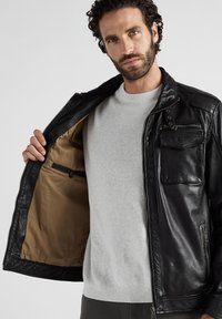 Schwarze Lederjacke mit Reißverschluss und mehreren Taschen. Innenfutter ist beige. Wird über einem hellgrauen Pullover getragen und zeigt einen lässigen Stil.