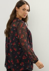 Kvinna med långt brunt hår klädd i en svart genomskinlig blus med blommönster i rött och grönt, som ser över sin axel mot en enkel bakgrund.