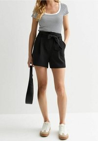 Shorts noirs taille haute avec un détail de nœud à la taille, associés à un haut ajusté rayé blanc et noir et des baskets blanches.