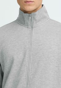 Veste polaire grise zippée avec un col haut, présentant une texture douce et un accent côtelé le long de la fermeture éclair et des coutures.