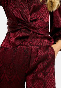 Bordová blúzka s textúrovaným, vzorovaným dizajnom, s uzlovým detailom v páse a elastickými manžetami na rukávoch.