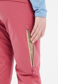 Pink waterproof pants with an angled zip pocket exposing a beige mesh interior. The zipper has a small purple pull tab. Smooth texture.