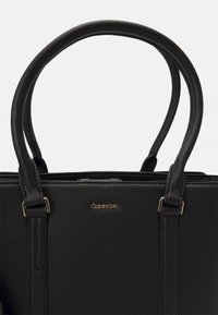 Czarna skórzana torebka z podwójnymi zaokrąglonymi uchwytami, teksturowaną powierzchnią, złotym logo oraz usztywnionym kształtem.