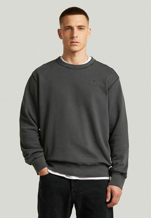 Giovane uomo con capelli corti che indossa una felpa girocollo grigio scuro sopra una camicia bianca, in piedi davanti a uno sfondo grigio chiaro uniforme.