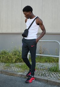 Sorte stribede bukser med rødt Nike-logo, hvid tanktop med lille blåt grafik, og sort crossbody taske; iført røde og sorte sneakers.