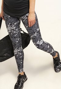 Legginsy w czarno-szarym wzorze kamuflażu, wykonane z elastycznego materiału, zestawione z czarnymi butami sportowymi i trzymające czarną torbę na siłownię.