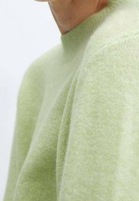Svetle zelený pletený svetr s vysokým výstřihem a jemnou texturou. Má uvolněný střih a subtilní žebrování na manžetách a spodním lemu.