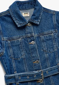 Denimmekko, jossa on napitushaitari, kaulus ja kaksi rintataskua. Varustettu vyötärölle sidottavalla vyöllä ja klassisen sinisellä värillä, jossa on oranssia ommelta.