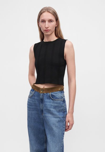 Jeune femme portant un crop top noir sans manches et un jean bleu taille haute avec une ceinture marron, sur un fond clair uni.