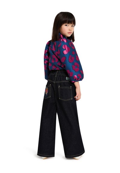 Un enfant porte un chemisier en satin bleu avec des motifs de cœurs roses et un pantalon en denim foncé à jambes larges avec des fleurs brodées sur les poches arrière.