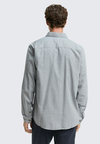 Uomo con capelli ricci castani, che indossa una camicia a maniche lunghe grigia a scacchi e pantaloni scuri, in piedi di spalle contro uno sfondo chiaro.