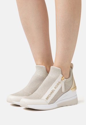 Sneaker slip-on in maglia beige con cerniera e rifiniture dorate. Presenta una superficie texturizzata e una suola a zeppa, etichettata "MICHAEL KORS."