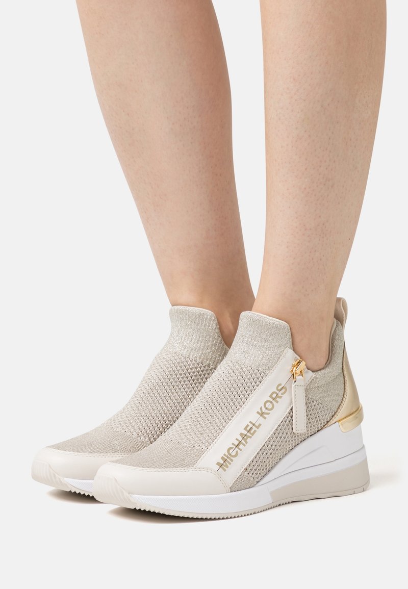 Sneaker slip-on in maglia beige con cerniera e rifiniture dorate. Presenta una superficie texturizzata e una suola a zeppa, etichettata "MICHAEL KORS."