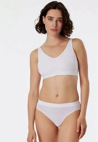 Woman with short curly hair wearing a white seamless bra and matching white underwear, standing against a plain light background.