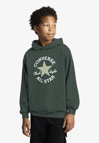 Žalias džemperis su gobtuvu, pagamintas iš minkšto medvilnės audinio, su kišene priekyje ir baltu žvaigždės logotipu bei „Converse All Star“ tekstu apskritimo forma.