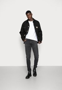 Black cotton jacket with a buttoned front and a logo patch, paired with a white t-shirt and dark grey slim-fit jeans, completed with black boots.