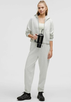 Sudadera gris clara con cordones y bolsillos, combinada con joggers a juego. Botella de agua negra con popote que presenta el nombre de la marca.