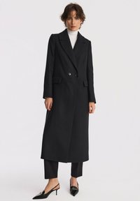Czarny wełniany płaszcz o długiej, prostej sylwetce, podwójnie zapięty, z szerokimi klapami i dwoma przednimi kieszeniami. Noszony na czarnych spodniach i na wysokich obcasach.