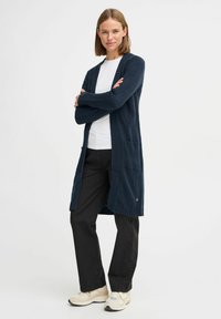 Marinblå stickad kofta med öppen front, långa ärmar och fickor. Stylerad över en vit t-shirt och svarta byxor, med ribbade muddar.