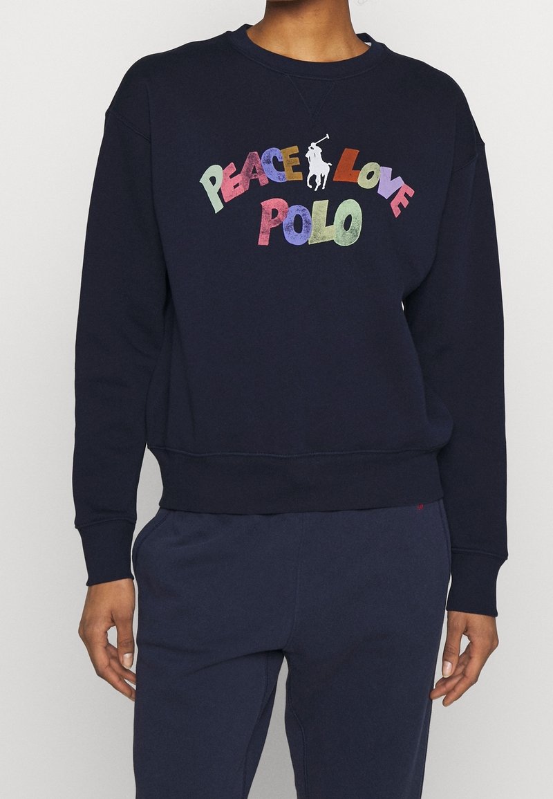 Personne portant un sweat-shirt bleu marine avec un texte coloré « PEACE LOVE POLO » et un logo blanc de joueur de polo sur la poitrine.