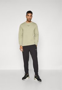 Lys olivengrøn sweatshirt med runde halsudskæring og sort logo, parret med mørke, tætsiddende sweatpants. Sorte træningssko med grønne accenter.