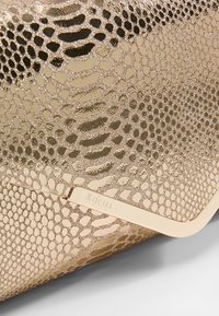 Złota kopertówka o teksturowanym wzorze wężowej skóry i metalicznym wykończeniu. Posiada metalowy detal z wygrawerowanym napisem "MASCARA". Gładki, elegancki design.