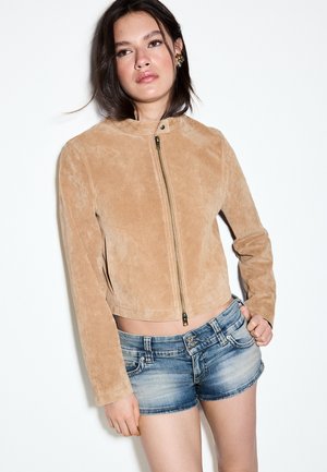 Jeune femme aux longs cheveux foncés portant une veste en daim beige avec une fermeture éclair à l'avant et un short en jean bleu, se tenant devant un fond blanc.