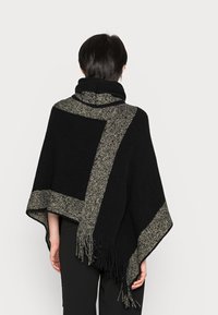 Poncho noir avec une bordure tissée contrastante en or et argent, présentant des bords à franges. Texture douce, coupe décontractée et design à capuche.