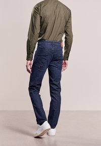 Man i mörkblå jeans, olivgrön långärmad tröja och vita sneakers, sedd bakifrån mot en enfärgad golv- och väggbakgrund.