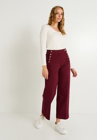 Pantalones anchos burdeos con cintura alta y cuatro detalles de botones metálicos en el costado. Combinados con una camiseta blanca de manga larga y zapatillas blancas.