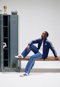 Αθλητική ζακέτα ναυτικού με λευκές ρίγες, φαρδιά μπλε τζιν με ρίγες και λευκά παπούτσια Adidas. Γκρι ντουλάπι στο φόντο με αθλητικά αντικείμενα.