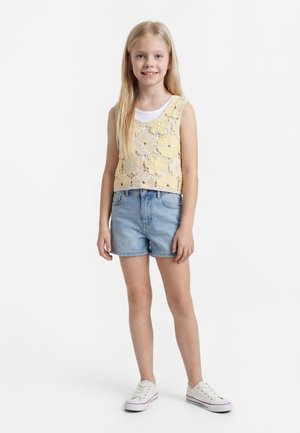 Ung jente står, iført en gul blomstrete heklet ermeløs topp over en hvit skjorte, denimshorts og hvite joggesko på hvit bakgrunn.