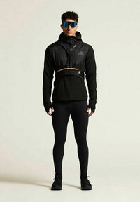 Schwarze Sportjacke mit Kapuze und Handschuhen, enganliegende schwarze Hose und blaue Sonnenbrille. Verfügt über ein Geschirr mit orangefarbenen Akzenten und ein elegantes Design.