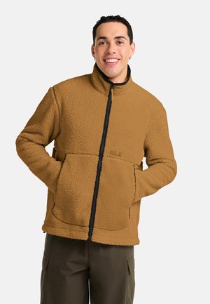 Junger Mann lächelt mit den Händen in den Taschen, trägt eine braune Fleecejacke und dunkle Hosen vor einem einfarbigen hellen Hintergrund.