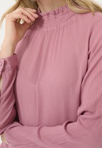 Blusa a maniche lunghe in morbido tessuto rosa con superficie testurizzata, dotata di un colletto alto con ruche e polsini bottonati per una vestibilità regolabile.