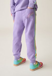 Lila Jogginghosen mit Regenbogen-Seitenstreifen und einer regenbogenbestickten Gesäßtasche, kombiniert mit mehrfarbigen Turnschuhen.