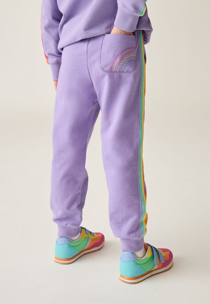 Lila Jogginghosen mit Regenbogen-Seitenstreifen und einer regenbogenbestickten Gesäßtasche, kombiniert mit mehrfarbigen Turnschuhen.