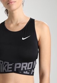 Žena v černém sportovním podprsenkovém topu Nike s bílým logem swoosh a nápisem "NIKE PRO" na elastickém pásu, na jednotném šedém pozadí.
