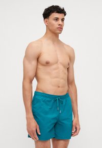 Shorts de bain en teal avec une taille élastique et un cordon de serrage. Le tissu semble lisse, avec un design minimaliste et sans motifs ni logos visibles.