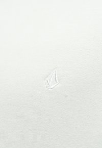 Tissu en coton blanc comportant un petit logo brodé blanc en forme de feuille, placé au centre.