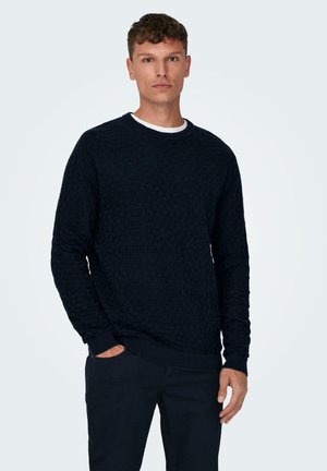 Pull en maille bleu marine avec un motif géométrique, col rond, poignets et ourlet côtelés, porté sur un pantalon sombre, mains dans les poches.