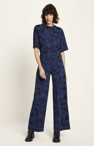 Woman with short dark hair stands wearing a blue and black patterned jumpsuit and black ankle boots against a plain background.