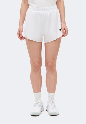 Női lábak fehér sportos rövidnadrágban, fehér zokniban és fehér futócipőben, világos egyszínű háttér előtt állva.