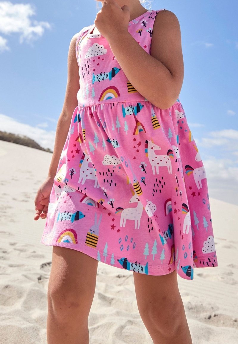 Enfant portant une robe rose avec des motifs de licorne, arc-en-ciel, chien et nuage, debout sur un terrain sablonneux sous un ciel bleu éclatant.