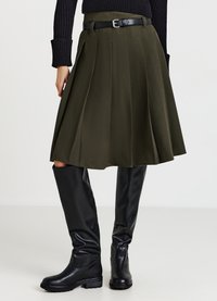 Gonna plissettata verde oliva con cintura nera; abbinata a stivali neri al ginocchio. Tessuto liscio, vita aderente, orlo svasato.