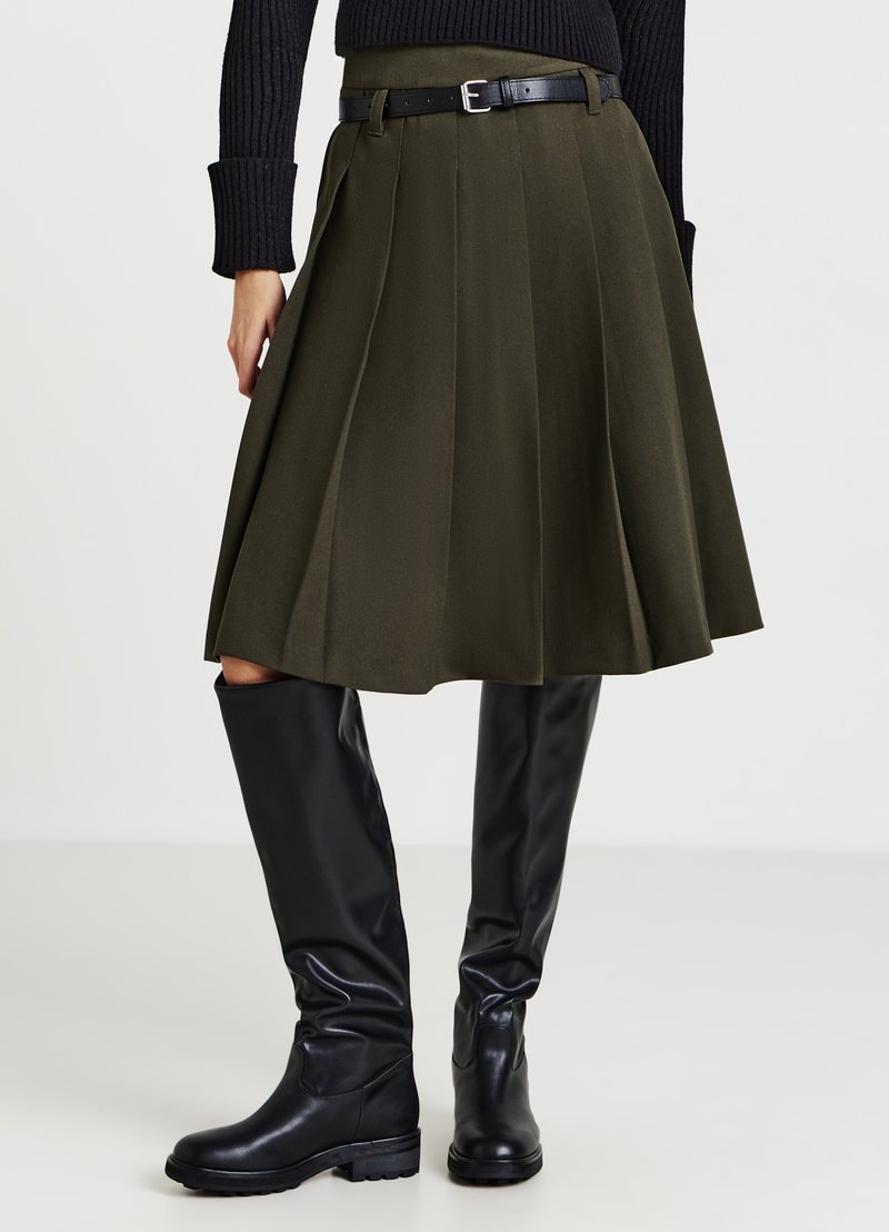 Gonna plissettata verde oliva con cintura nera; abbinata a stivali neri al ginocchio. Tessuto liscio, vita aderente, orlo svasato.